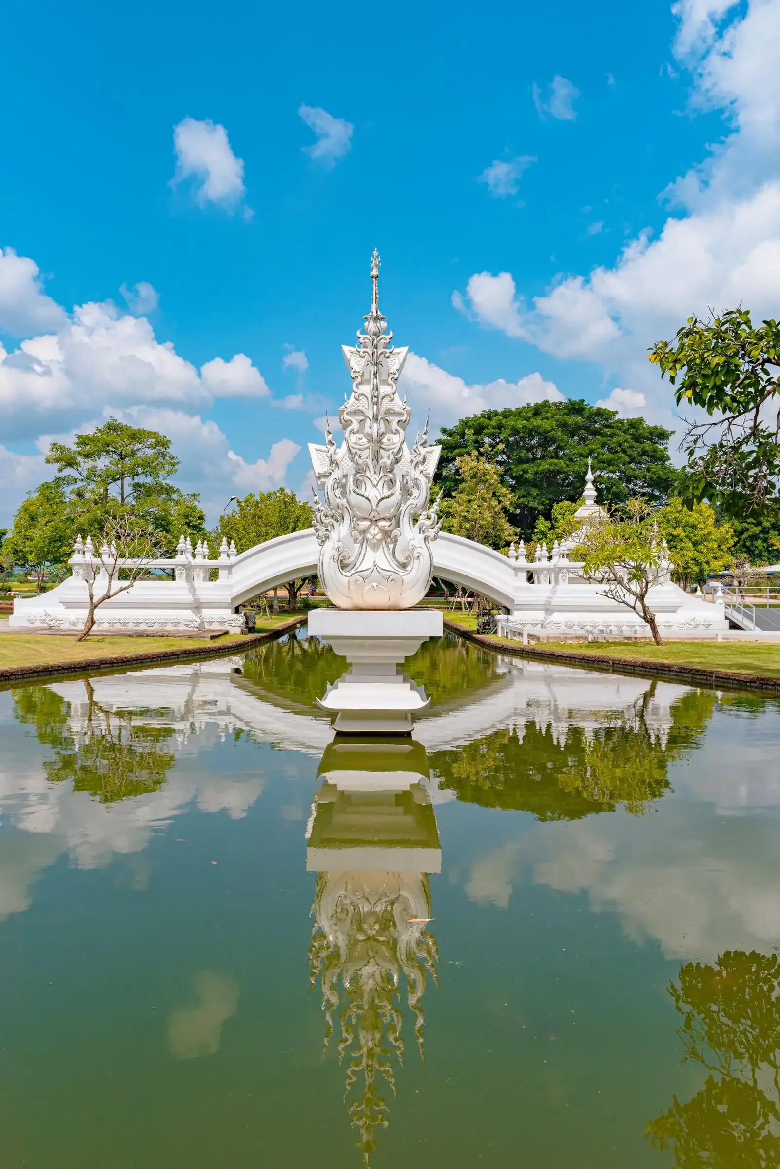 A white spire in a pond between white arched bridges at Wat Rong Khun