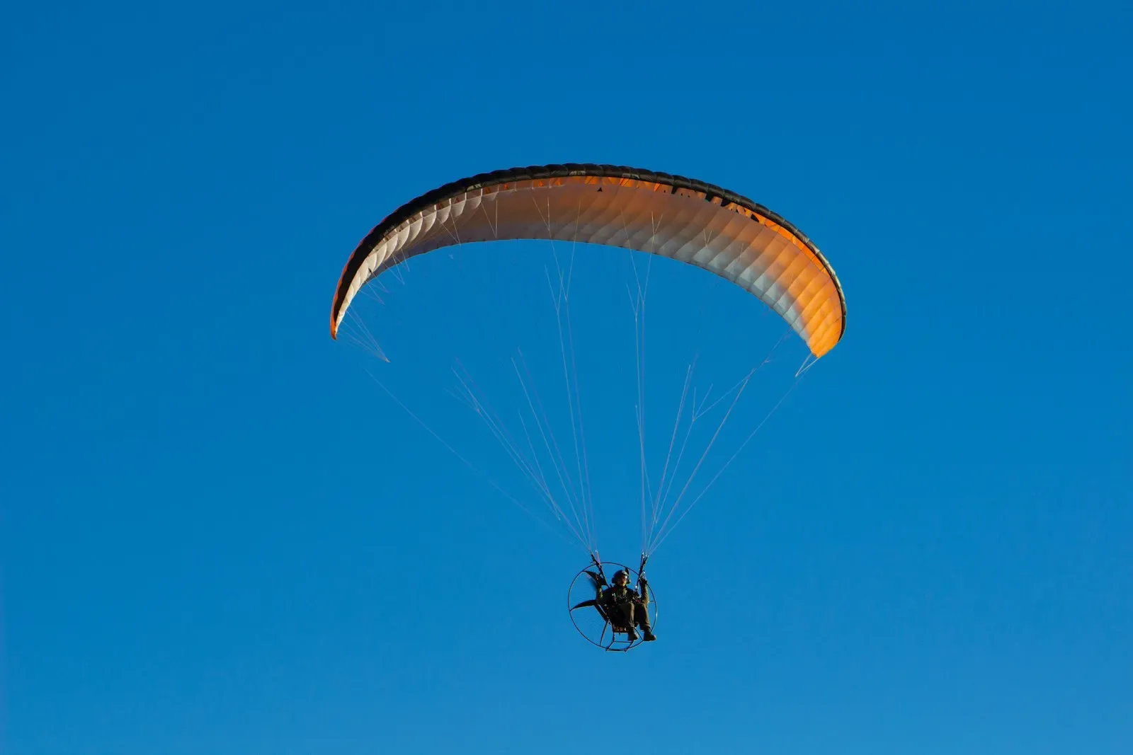 A paramotor pilot under an orange wing against a blue sky